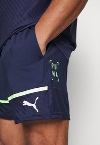 Mies, joka pukeutuu tummansiniset Puma-urheilushortsit neonvihreillä yksityiskohdilla, käsi oikeassa taskussa, näkyvä jalka ja käsivarsien läheltä otettu kuva.