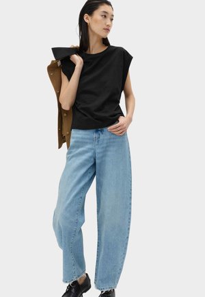 Kvinde iført sort ærmeløs top, lyseblå vidde ben jeans, sorte sko, holder en brun jakke over skulderen, står mod hvid baggrund.