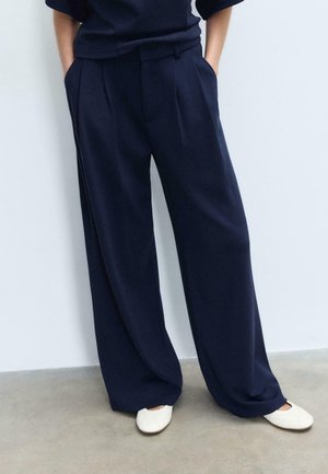 Osoba nosząca luźne granatowe spodnie i białe płaskie buty, stojąca z rękami w kieszeniach na tle gładkiej ściany.