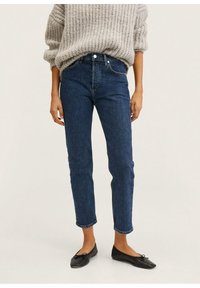 Jean en denim foncé taille haute avec une jambe fuselée, doté d'une fermeture à boutons sur le devant et de détails de coutures minimales. Porté avec un pull en tricot gris clair.