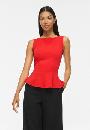 Femme portant un haut rouge sans manches à basque et un pantalon noir à jambes larges, debout avec une main dans la poche sur fond blanc.