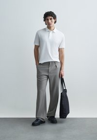 White short-sleeve polo shirt, gray pleated trousers, black sneakers, and a black tote bag. Smooth fabric, fitted design, and neutral colors.