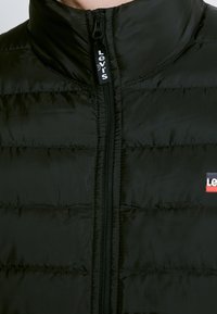 Veste matelassée noire avec col droit, fermeture éclair centrale avec tirette à logo Levi's et patch partiel du logo Levi's sur la poitrine.