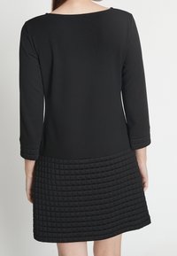 Svart klänning med en slät överdel och en texturerad, vadderad nederdel. Har trekvartslånga ärmar och en rundad halsringning.