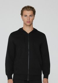 Schwarze Zip-Hoodie aus Baumwolle. Verfügt über eine Kapuze mit Kordelzug, Fronttaschen und gerippte Bündchen sowie einen gerippten Saum. Glatte Textur und enganliegendes Design.