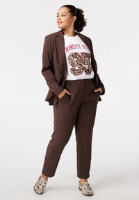 Costume marron ajusté avec un t-shirt graphique blanc arborant un design léopard "95". Chaussures à enfiler avec une surface texturée et décorée.