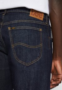 Dark blue Lee brand jeans with yellow stitching worn by a person with dark skin, visible hand by their side, and a white shirt tucked in.