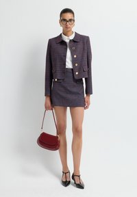Giacca di tweed su misura in blu navy e bordeaux con bottoni dorati, abbinata a una mini gonna coordinata. Gli accessori includono una borsa rossa e tacchi neri.