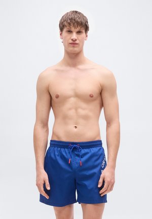 Blue swim shorts with a drawstring waist, red accents, and a printed logo. Made from lightweight, smooth fabric.