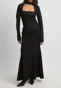 Schwarzes Langarmoberteil mit quadratischem Ausschnitt und taillierter Passform, kombiniert mit einem bodenlangen schwarzen Rock und spitzen schwarzen Absätzen.