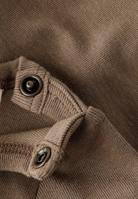 Gros plan sur un tissu brun doux avec des coutures visibles et deux boutons noirs, mettant en valeur la texture et les détails de design.