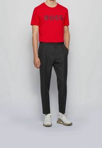 Mand iført rød T-shirt med teksten "BOSS", mørkegrå skræddersyede bukser og hvide sneakers, stående mod en ensfarvet lys baggrund.