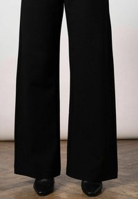 Zwarte wijde broek gemaakt van een gladde, zachte stof. Heeft een hoge taille met minimale naden. Gdragen met zwarte schoenen op een houten vloer.