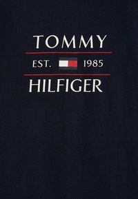 Námořnická modrá bavlněná tričko s nápisem "TOMMY" a "HILFIGER" v bílé barvě, s "EST. 1985" a malým logem v červené, bílé a modré barvě.