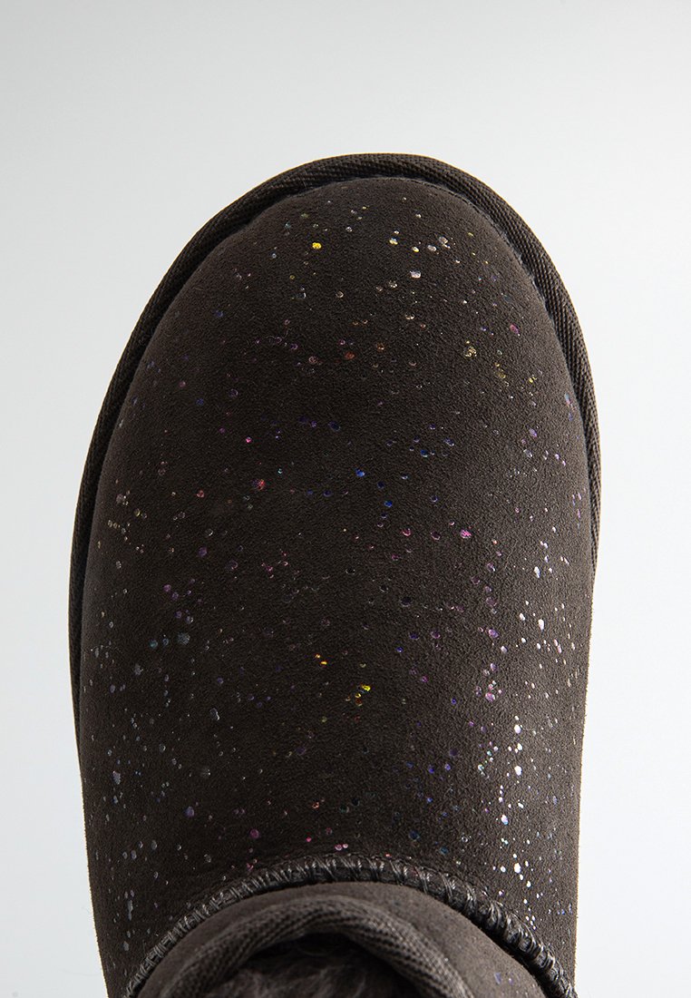 Zwarte suède schoen met multicolore glitters en gestikte bies langs de afgeronde teen tegen een effen lichte achtergrond.