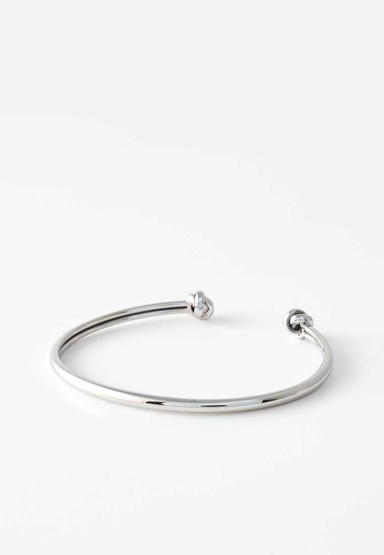 Bracciale in metallo argenteo a forma di manicotto, caratterizzato da una forma liscia e arrotondata. Presenta due estremità piccole e arrotondate e una superficie lucida e riflettente.