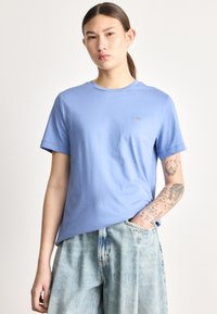 Svetlo modra bombažna majica s kratkimi rokavi in okroglim izrezom. Nežen logotip na prsih. Oblečena s posvetljenimi jeansi kratkimi hlačami.