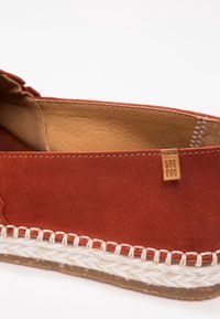Chaussure à enfiler en daim rouge avec une doublure en cuir naturel, accentuée par des coutures blanches tressées et une petite étiquette sur le côté.