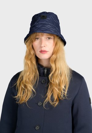 Marineblauwe gewatteerde bucket hat van glanzend materiaal, met een ronde logopatch. Draagt in combinatie met een bijpassende marineblauwe jas met een ritssluiting bij de kraag.