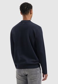 Marineblauwe polo met lange mouwen en een textuur gebreid oppervlak, voorzien van een kraag en ribgebreide manchetten. Grijze spijkerbroek zichtbaar aan de onderkant.
