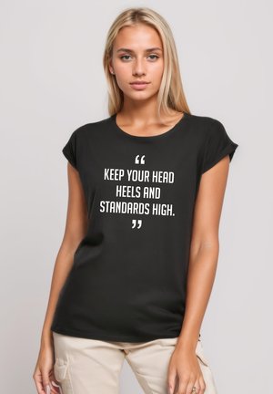 Schwarzes T-Shirt mit kurzen Ärmeln aus Baumwolle, das mit weißem Text bedruckt ist, der lautet: "HALTE DEINEN KOPF, DEINE FÜSSE UND DEINE ANSPRÜCHE HOCH." Klassisches Rundhalsdesign.