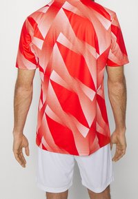 Jersey deportivo rojo con un patrón gráfico de formas angulares en blanco y rojo claro. Manga corta y tejido ligero. Pantalones cortos blancos debajo.