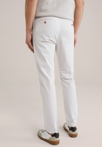 Pantaloni di cotone bianchi con una vestibilità slim, dotati di due tasche posteriori con bottoni marroni e una texture liscia, abbinati a scarpe da ginnastica casual.