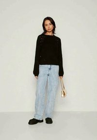 Sweater preto canelado, jeans largos azul claro, sapatos pretos robustos e uma pequena mala dourada. Modelo de pé contra um fundo neutro.