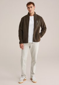 Chaqueta de forro polar marrón con un bolsillo en el pecho con cremallera, cuello alzado y textura suave, llevada sobre una camisa blanca y pantalones claros.