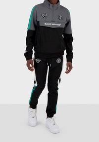 Svart träningsdräkt med grå detaljer, som har en halvdragkedja jacka och logotypapplikationer. Byxorna har sidostripes och vita sneakers.