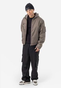 Hooded bomber jacket in taupe, featuring elastic cuffs and a zippered pocket. Worn over a black t-shirt with loose black cargo pants and sneakers.