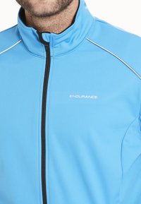 Lysblå atletisk jakke med høj krave, tynd sort lynlås og hvid accentsyning. Har "ENDURANCE" logoet på brystet.