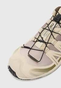 Beżowe buty do biegania w terenie z siateczkową cholewką, grubą podeszwą i elastycznymi sznurowadłami. Szczególne detale to teksturowane wykończenie oraz akcenty z logo.