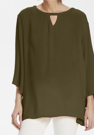 Luźna bluzka w oliwkowej zieleni z rękawami 3/4 i małym wycięciem w kształcie łezki poniżej okrągłego dekoltu, noszona z białymi spodniami.