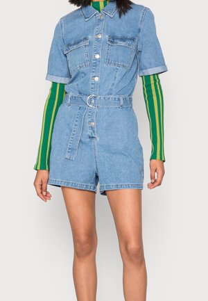 Vrouw draagt een spijkerromper met korte mouwen en ceintuur over een groen-gele gestreepte top met lange mouwen, staand tegen een effen lichte achtergrond.