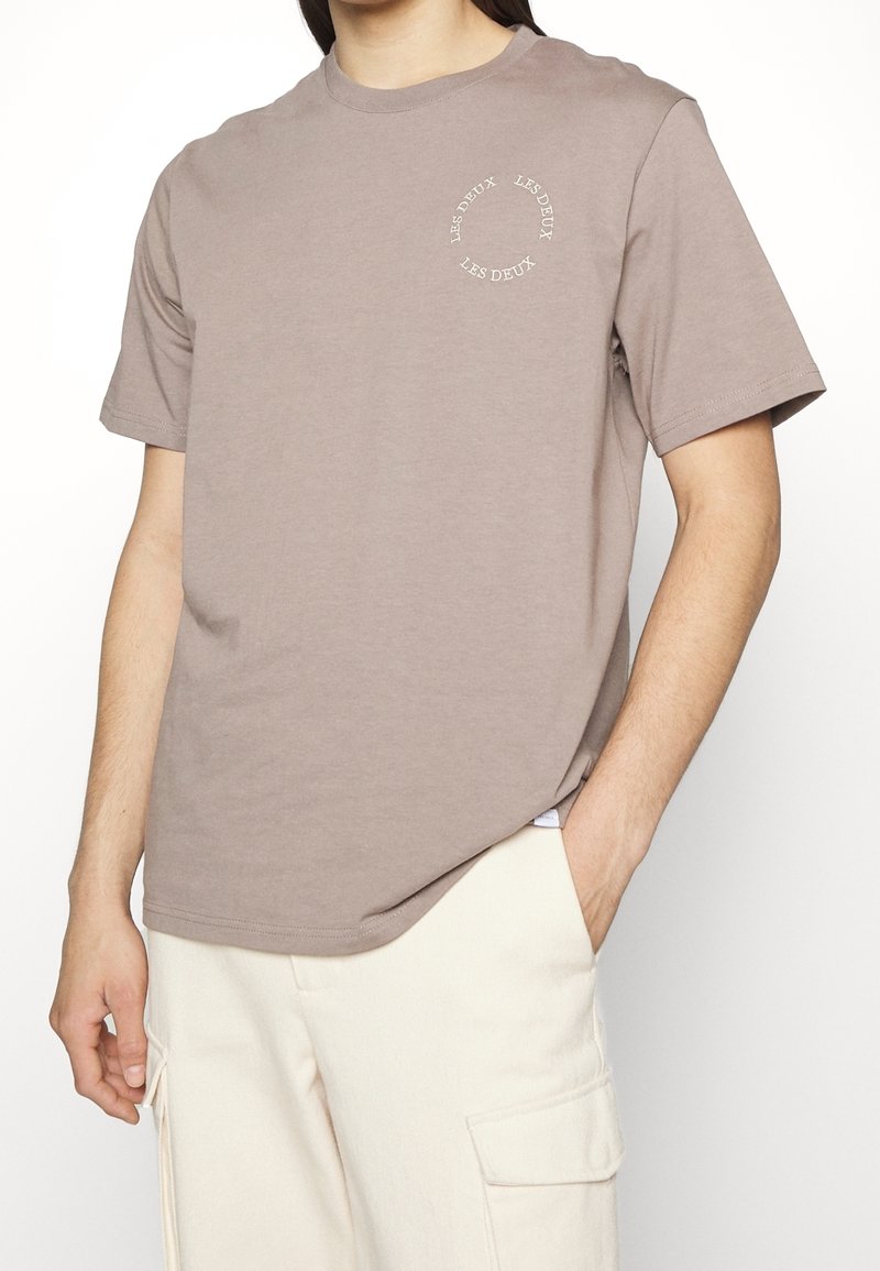 Tričko s krátkým rukávem a kulatým výstřihem v tlumené taupe barvě, vyrobené z měkké bavlny. Na hrudi se nachází kruhový bílý logotyp s textem, standardní střih.