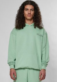 Šviesiai žalias gobtuvėlis, pagamintas iš minkštos medžiagos, su priekinėmis kišenėmis, raišteliu ant gobtuvo ir išsiuvinėtu "Garbingas ir Šlovingas" užrašu ant kairiojo krūtinės.