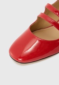 Roter Lacklederschuh mit einer abgerundeten Spitze und drei dekorativen Riemen über dem Spann. Glatte Oberfläche und eine flache schwarze Sohle.