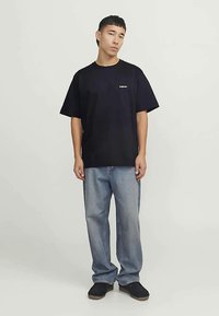 Jaunas vyras stovi atsipalaidavęs, vilkėdamas juodą per didelį marškinį, laisvas mėlynas džinsus ir juodas batus prieš vienodą fono.