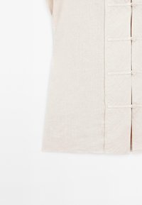 Beżowa lniana tunika o prostym kroju, z krótkimi rękawami i zapięciem z przodu na materiałowe pętelki i guziki. Gładka faktura.