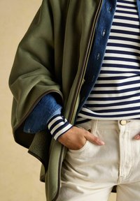 Person trägt geschichtete Kleidung: eine grüne Fleecejacke, ein blaues Jeanshemd, ein marineblau-weiß gestreiftes Hemd und helle Hose, eine Hand in der Tasche.