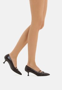 Scarpe con tacco nere in pelle con punta affusolata, caratterizzate da cinturini e anelli decorativi, e un tacco piccolo. Texture liscia e lucida.