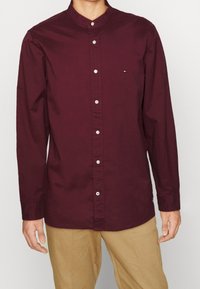 Camisa de manga larga burdeos con cuello de banda, con botones blancos y un pequeño logotipo en el pecho izquierdo. Tela de algodón, textura suave.
