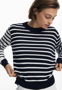 Marineblau-weiß gestreifter Pullover mit Rundhalsausschnitt. Ribbedetails an den Bündchen und dem Saum. Das Model hält den unteren Saum mit beiden Händen.