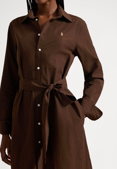 Woman wearing a brown button-up dress with long sleeves and a tied belt at the waist, showing Polo Ralph Lauren logo on chest.
