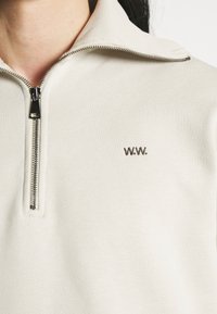 Světlá béžová mikina na zip s žebrovanou texturou, vysokým límcem a vyšívaným logem "W.W." na levé straně. Stříbrný detail zipu.