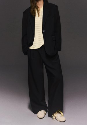 Person iført oversize sort blazer og bukser med vide ben, creme-stribet t-shirt og off-white sneakers med gule snørebånd, der står mod en mørk baggrund.