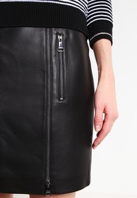 Jupe en cuir noir avec une texture lisse, dotée d'une fermeture éclair sur le côté et de détails en métal argenté, associée à un pull rayé noir et blanc.