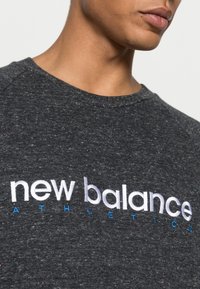 Camiseta gris texturizada con motas multicolores. Presenta un logo bordado de "new balance" en blanco y azul. Diseño de cuello redondo.