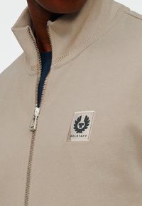 Άτομο που φοράει μπεζ μπουφάν με φερμουάρ και ψηλό γιακά, με το λογότυπο της Belstaff στο στήθος, πάνω από ένα σκουρομπλε πουκάμισο.